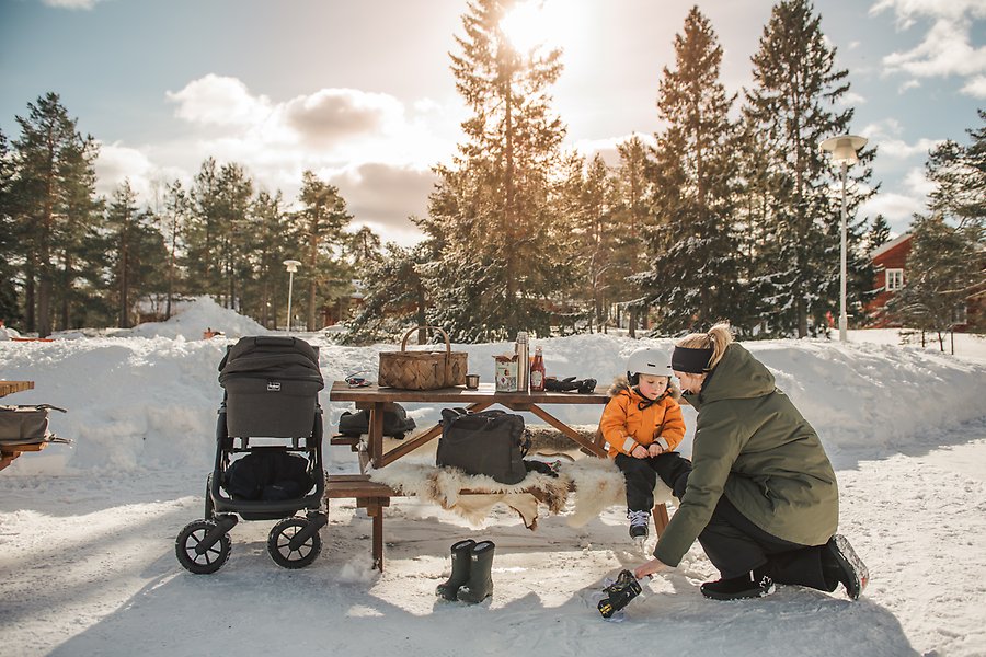 En mamma hjälper sitt barn att sätta på skridskor. De sitter på en bänk utomhus. Det är snö runtomkring och solen skiner.